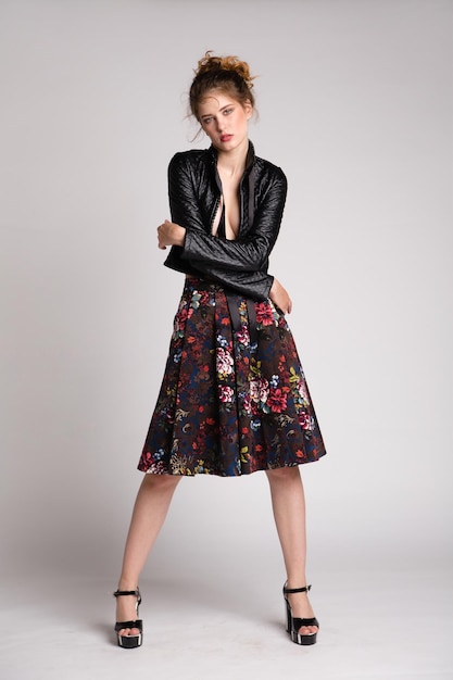 mujer joven elegante en falda larga de patrón floral negro, chaqueta de cuero posando sobre fondo blanco