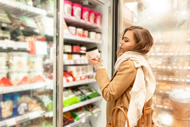 Mujer joven elige helado en la tienda. Una hermosa rubia sonriente con un abrigo beige abrió la puerta del refrigerador. Comida sana y sabrosa.