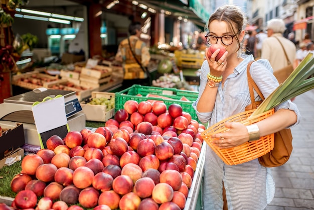 Mujer joven eligiendo un durazno fresco de pie con el canasto en el mercado de alimentos en Francia