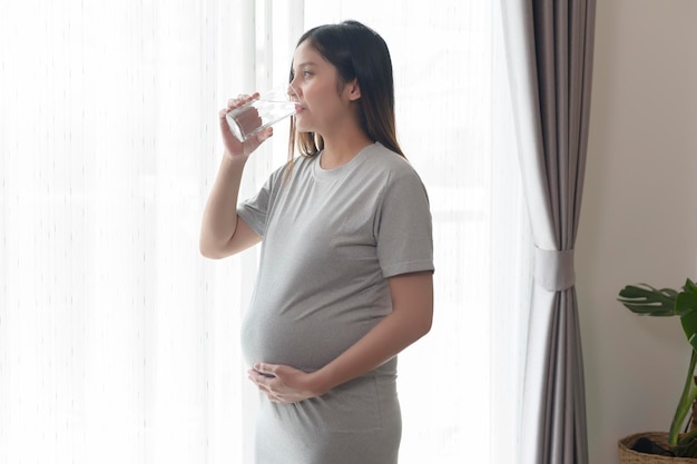 Mujer joven embarazada bebiendo agua en el hogar, atención médica y concepto de atención del embarazo