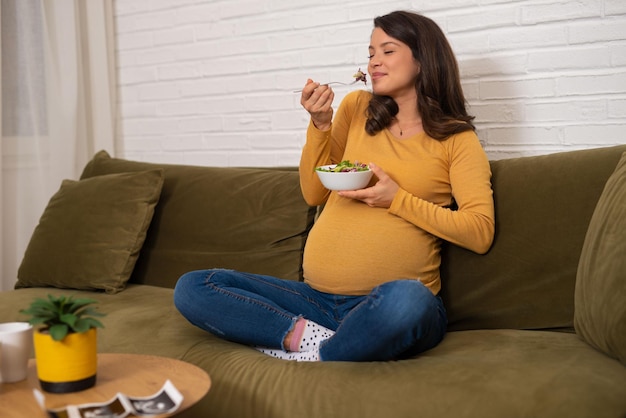 Mujer joven embarazada feliz sentada y comiendo ensalada de frutas en el sofá en casa