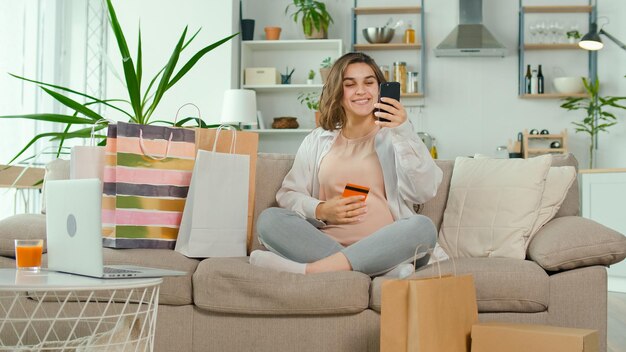 Mujer joven embarazada hace compras en línea usando su teléfono y tarjeta de crédito