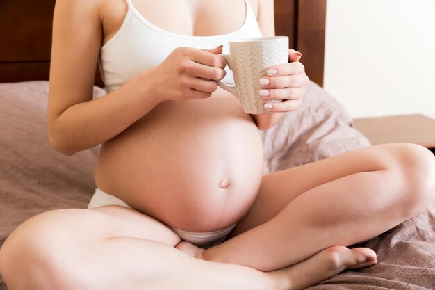 Foto mujer joven embarazada sentada en la cama y bebiendo café en casa concepto de maternidad