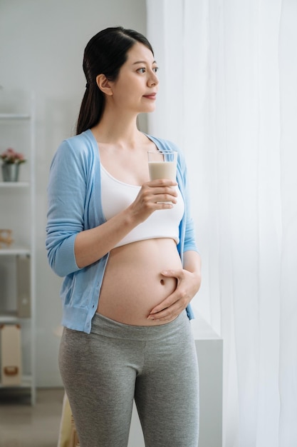 Mujer joven embarazada con vaso de leche de pie en la sala de estar. encantadora dama sonriente paternidad por la ventana mirando hacia afuera disfrutar del tiempo de descanso en casa relajarse solo. atractivo embarazo femenino asiático