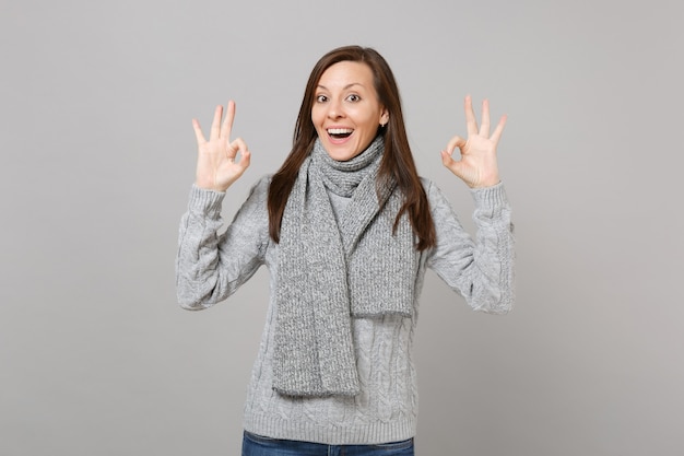 Mujer joven emocionada alegre en suéter gris, bufanda que muestra gesto OK aislado sobre fondo gris en estudio. Estilo de vida de moda saludable, personas emociones sinceras, concepto de estación fría. Simulacros de espacio de copia.