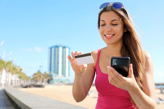 Mujer joven emocionada comprando en línea con teléfono móvil leyendo el número de tarjeta de crédito al aire libre en verano