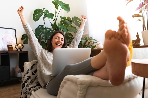 Mujer joven emocionada y feliz levantando los brazos para celebrar el éxito o el logro en casa usando una computadora portátil