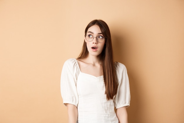 Mujer joven emocionada con gafas mirando a un lado con la boca abierta, jadeando fascinada, de pie sobre beige.