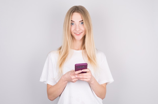 Mujer joven emocionada mirando con sorpresa a la cámara con teléfono móvil, sonriendo. Mujer leyendo un mensaje de texto en su teléfono, aislado sobre fondo blanco.