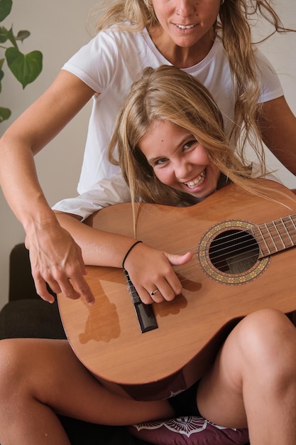 Mujer joven emocionada y positiva con una sonrisa dentada sentada en el sofá y tocando la guitarra acústica de madera mientras mira a la cámara