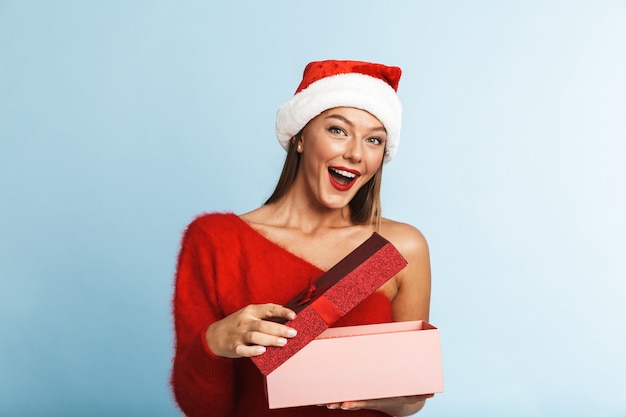 Mujer joven emocionada con sombrero rojo de santa claus de pie con un cuadro de presente abierto