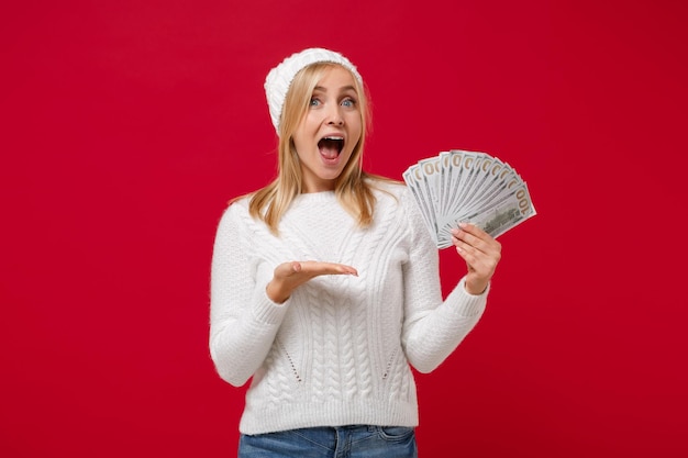 Mujer joven emocionada con suéter blanco, sombrero aislado en el fondo de la pared roja. Estilo de vida de moda saludable, concepto de temporada fría. Simulacros de espacio de copia. Apuntando con la mano al abanico de dinero en efectivo en billetes de dólar.