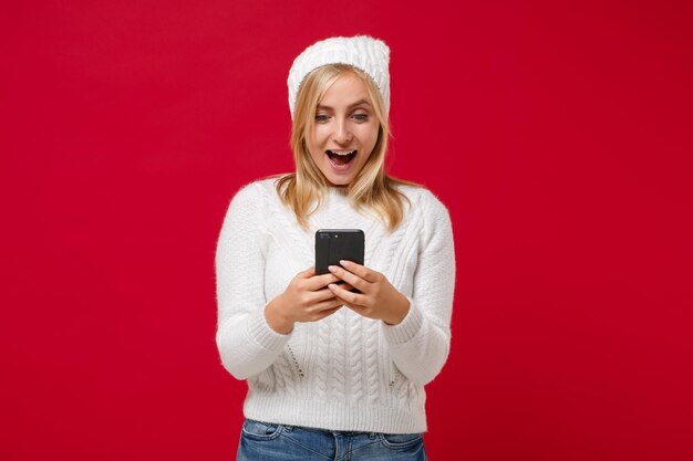 Mujer joven emocionada con suéter blanco, sombrero aislado en el fondo de la pared roja, retrato de estudio. Estilo de vida de moda saludable, concepto de temporada fría. Simulacros de espacio de copia. Usando teléfono móvil, escribiendo mensajes sms.