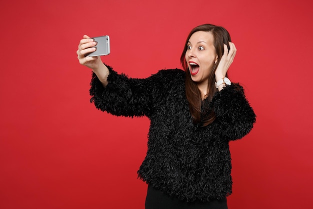 Mujer joven emocionada en suéter de piel negra aferrándose a la cabeza, haciendo selfie disparo en teléfono móvil aislado sobre fondo rojo brillante de la pared. Personas sinceras emociones, concepto de estilo de vida. Simulacros de espacio de copia.