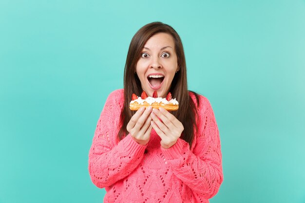 Mujer joven emocionada en suéter rosa tejido manteniendo la boca abierta mirando sorprendido, sosteniendo en las manos pastel de eclair aislado sobre fondo azul en estudio. Concepto de estilo de vida de personas. Simulacros de espacio de copia.