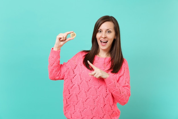 Mujer joven emocionada en suéter rosa tejido que señala el dedo índice en la moneda futura de bitcoin en la mano aislada sobre fondo de pared azul turquesa en estudio. Concepto de estilo de vida de personas. Simulacros de espacio de copia.