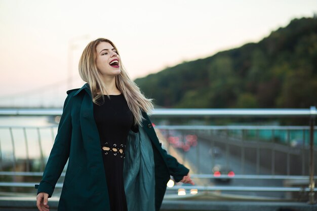 Mujer joven emocional con abrigo verde posando en el puente con luces. Espacio para texto