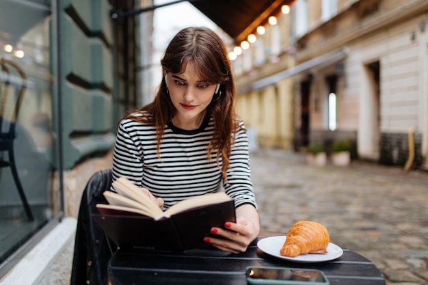 Mujer joven emocional en el libro de lectura de auriculares inalámbricos mientras está sentado en la acogedora terraza de un café