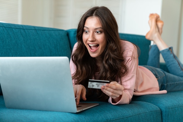 una mujer joven emocional sonriente adentro en casa usando la computadora portátil que sostiene la tarjeta de crédito en el sofá.