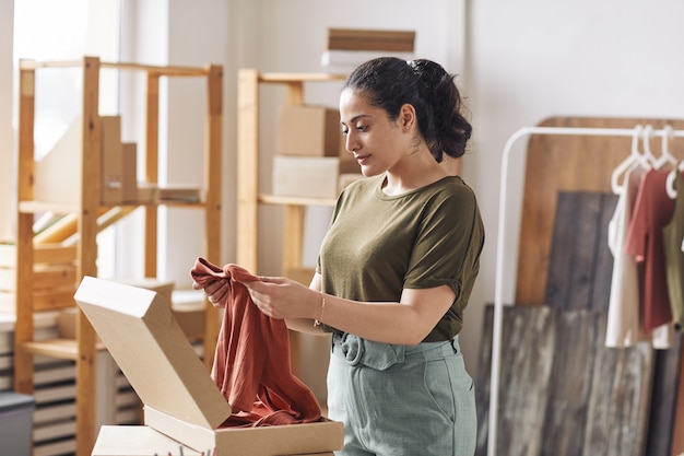 Mujer joven empacando camisa nueva en caja de cartón antes de entregar trabajando en tienda web