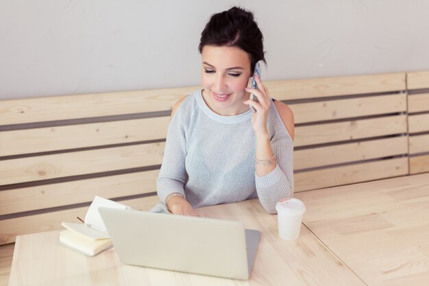 Mujer joven emprendedora casual que trabaja en línea escribiendo con una computadora portátil en la oficina o café y hablando con el teléfono celular