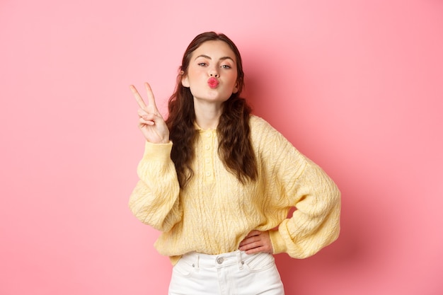 La mujer joven del encanto frunce los labios, haciendo la cara del beso y el signo v, muestra el gesto de la paz, de pie contra la pared rosada.