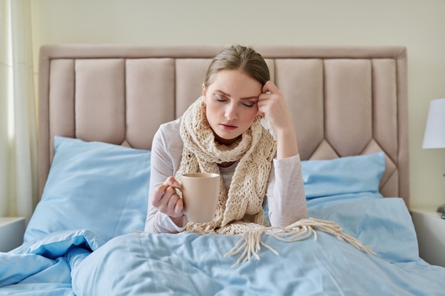 Mujer joven enferma en la cama con pañuelo y taza