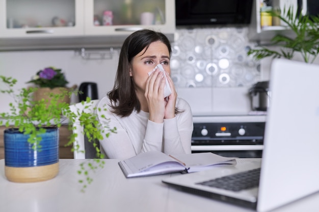 Mujer joven enferma. Estornudar y toser con pañuelo, sentado con el portátil en la oficina en casa, fondo interior de la cocina. Virus, resfriados estacionales, alergias, temporada de gripe