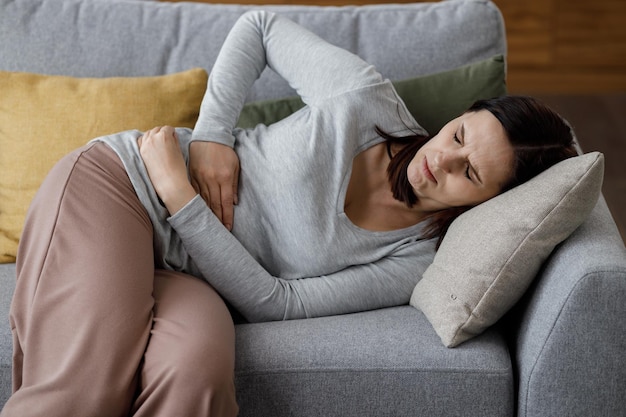 Mujer joven enferma que sufre de dolor menstrual Mujer con las manos apretando el vientre con dolor de estómago o calambres menstruales sentada en el sofá Dolor abdominal gastritis y concepto de períodos dolorosos