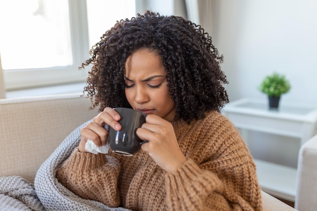 Mujer joven enferma sentada en casa en la cama con una taza de té caliente y un pañuelo Resfriados estacionales tos secreción nasal infecciones virales tratamiento en el hogar