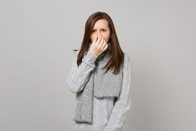 Mujer joven enferma en suéter gris, bufanda con secreción nasal, sosteniendo la mano en la nariz aislada sobre fondo de pared gris. Estilo de vida saludable tratamiento de enfermedades enfermas, concepto de estación fría. Simulacros de espacio de copia.