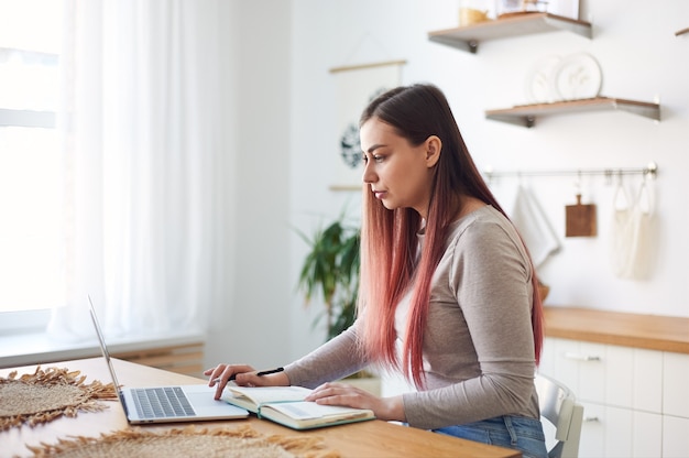 Mujer joven enfocada viendo un webinar, tomando notas en un cuaderno. Aprendizaje a distancia en casa. Trabajo remoto.