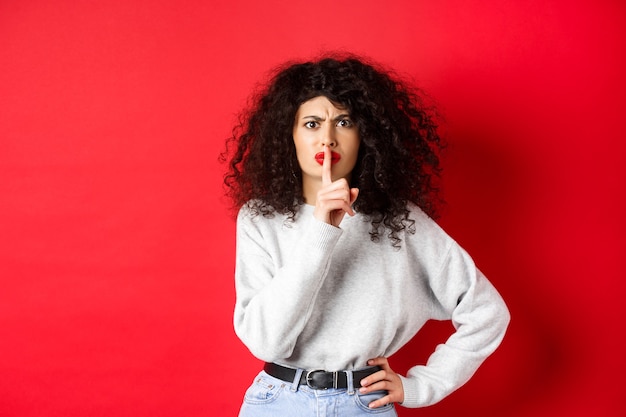 Mujer joven enojada con el pelo rizado y labios rojos frunciendo el ceño y callando dice que se calla hacer tabú ges ...