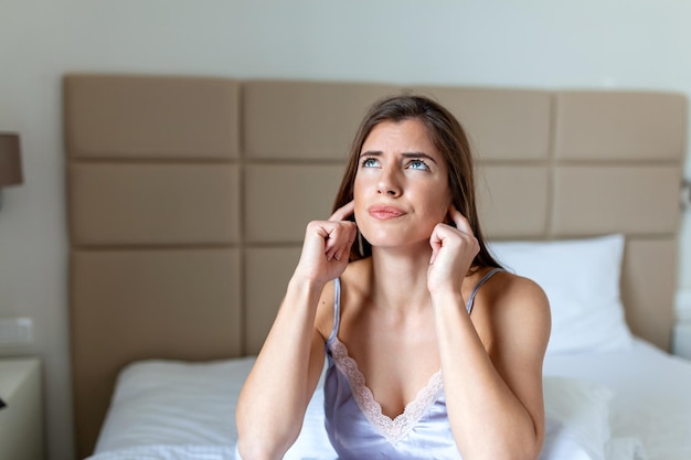 Mujer joven enojada que sufre y está perturbada por vecinos ruidosos y se cubre los oídos con los dedos mientras intenta dormirse en la cama en casa temprano en la mañana