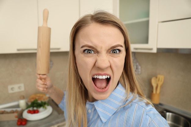 Mujer joven enojada con rodillo en la cocina