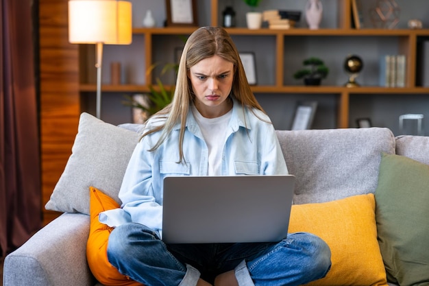 Mujer joven enojada sentada en el sofá en la sala de estar con problemas de funcionamiento de la computadora portátil molesta por la conexión lenta a Internet chica confundida que trabaja desde casa experiencia falla del software del sistema