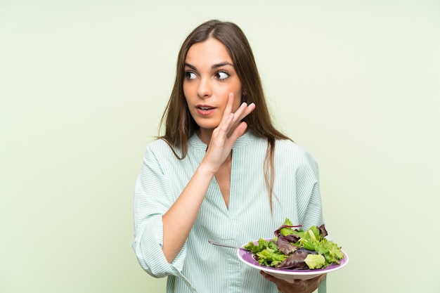 Foto mujer joven con ensalada sobre pared verde aislada susurrando algo