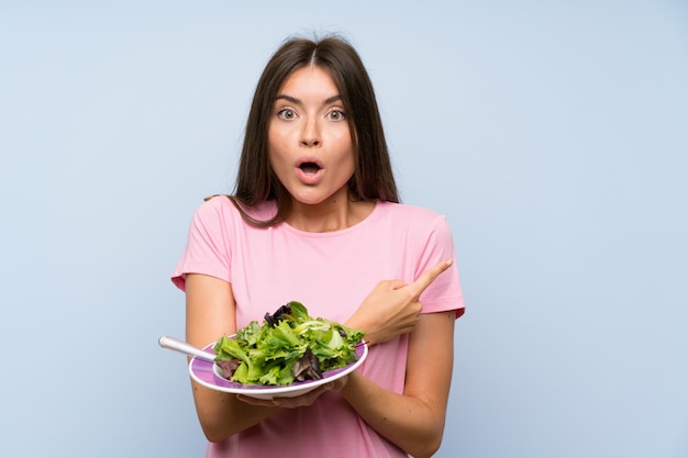 Mujer joven con ensalada sorprendida y apuntando hacia el lado