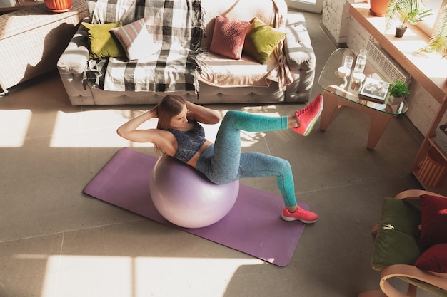 Foto mujer joven enseñando en casa cursos en línea de fitness, estilo de vida aeróbico y deportivo mientras está en cuarentena. ponerse activo mientras está aislado, bienestar, concepto de movimiento. ejercicios con fitball para la parte inferior del cuerpo.