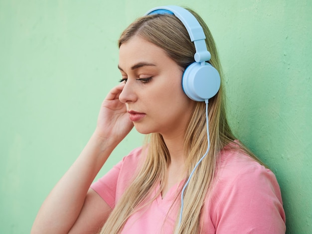 Mujer joven ensimismada en una camiseta rosa escuchando música en auriculares azules mientras está de pie contra una pared verde