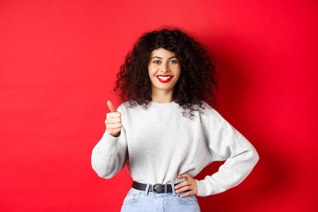 Mujer joven entusiasta con cabello rizado, labios rojos, mostrando los pulgares hacia arriba y sonriendo con aprobación, elogia el buen producto, de pie contra la pared roja