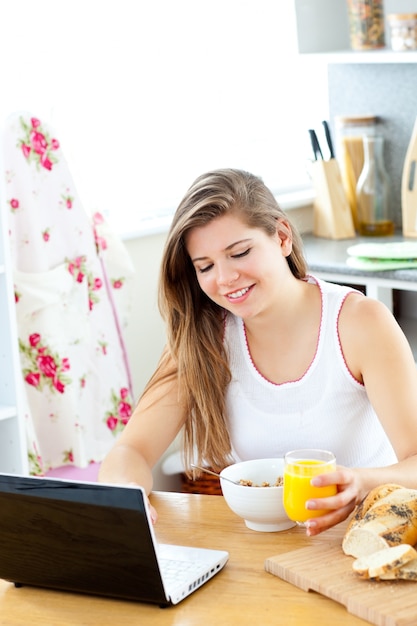 Mujer joven entusiasta que usa su computadora portátil mientras que desayuna en la cocina