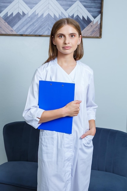 Una mujer joven es una médica interna que lleva una bata blanca de pie con una carpeta en las manos