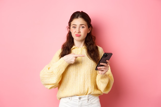 Mujer joven escéptica haciendo muecas insatisfecho con el dedo acusador en la pantalla del teléfono inteligente que tiene dudas sobre el contenido en línea de pie contra la pared rosa