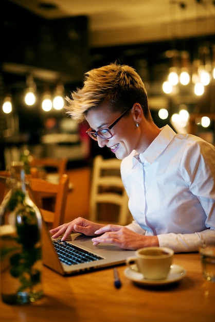 Mujer joven escribiendo en la computadora portátil en el café. Sonriendo con gafas mirando portátil.