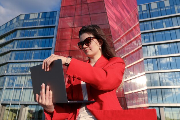 Mujer joven escribiendo en una computadora portátil en frente de un edificio de oficinas