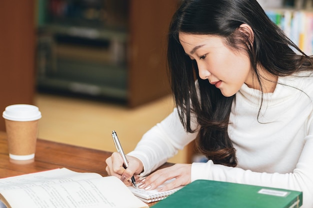 Mujer joven escribiendo en un libro en la mesa de la biblioteca