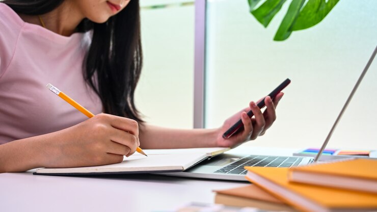 Foto mujer joven escribiendo en papel de cuaderno y leyendo en la pantalla del teléfono inteligente.