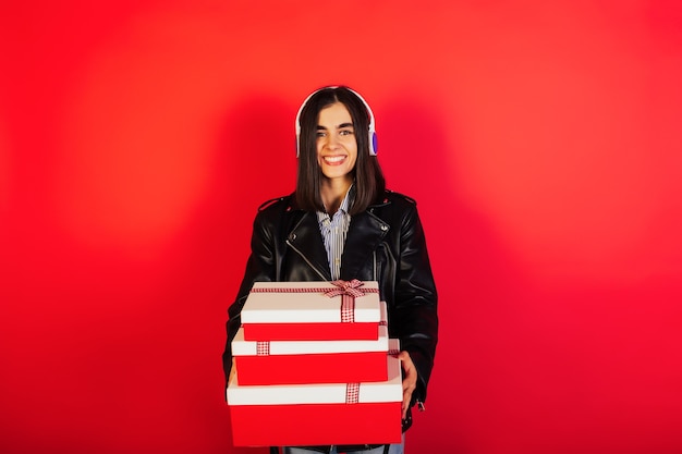 Mujer joven escucha música con auriculares y sosteniendo tres cajas de regalo en la pared roja