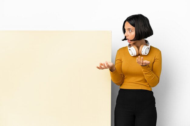 Mujer joven escuchando música con un gran cartel vacío sobre fondo aislado con expresión de sorpresa mientras mira de lado